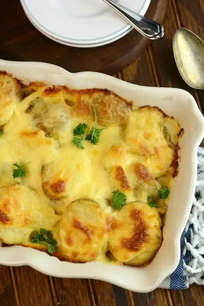 A baking dish with easy potatoes au gratin, topped with a cheesy crust and garnished with parsley, sits on a wooden table alongside a serving spoon and plates.