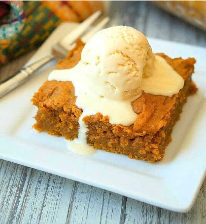 A slice of pumpkin pudding cake recipe is topped with a scoop of melting vanilla ice cream on a white plate.