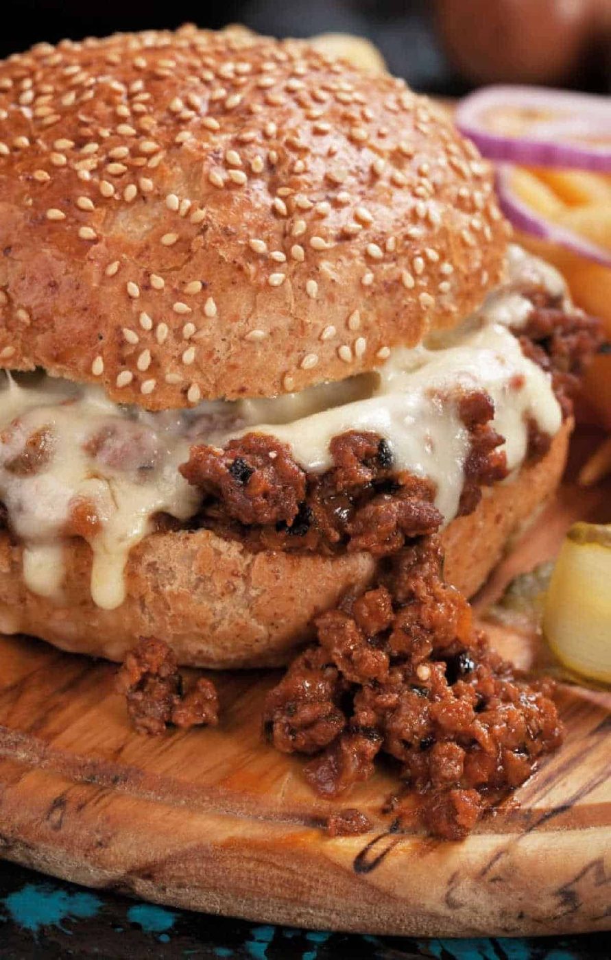 Close-up of a sloppy joe sandwich made with an easy sloppy joe recipe 3 ingredients, topped with melted cheese on a sesame bun, served on a wooden board.