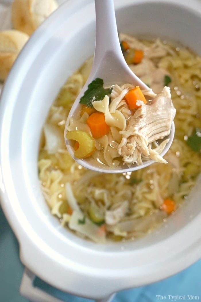 A ladle scoops chicken noodle soup from a white crockpot, revealing tender noodles, succulent chicken, vibrant carrots, and fresh herbs.