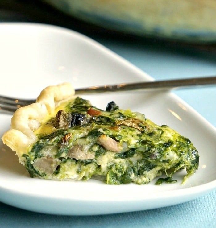 A slice of easy spinach quiche recipe featuring mushrooms rests on a white plate with a fork.