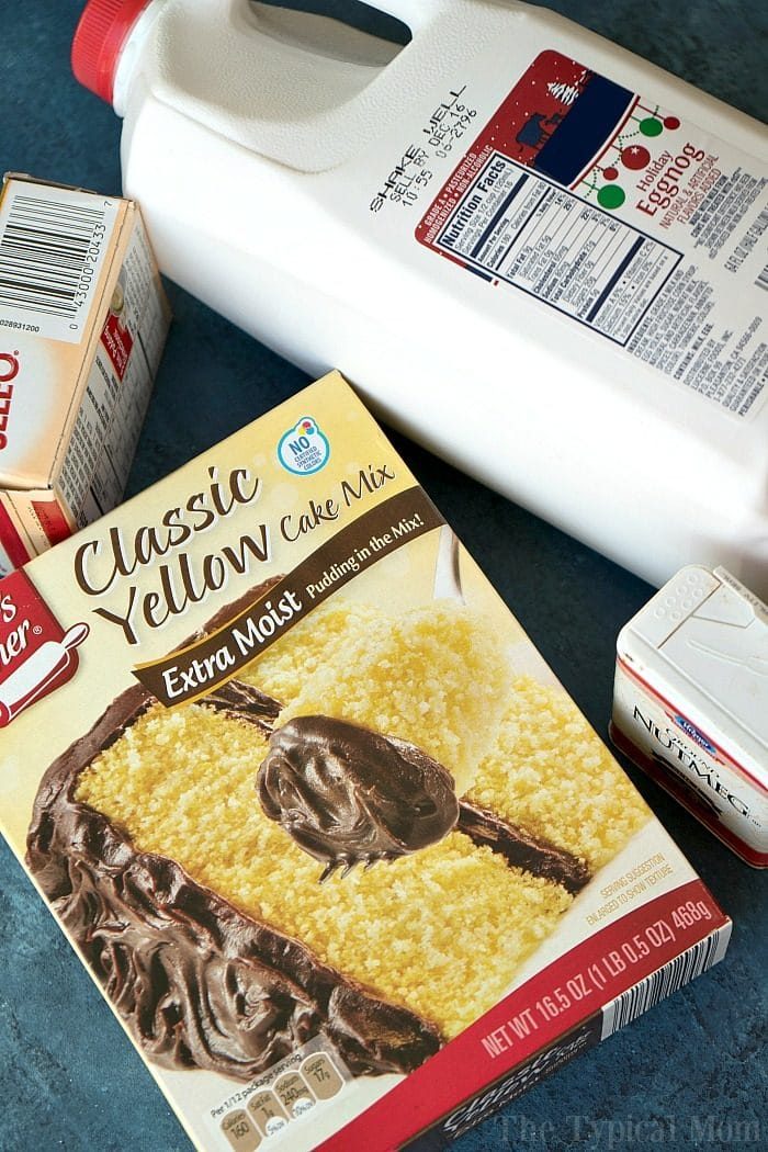 A box of classic yellow cake mix sits on a blue surface, accompanied by a carton of milk, and unsalted butter. The cake mix box showcases an image reminiscent of an egg nog pudding cake adorned with chocolate frosting.