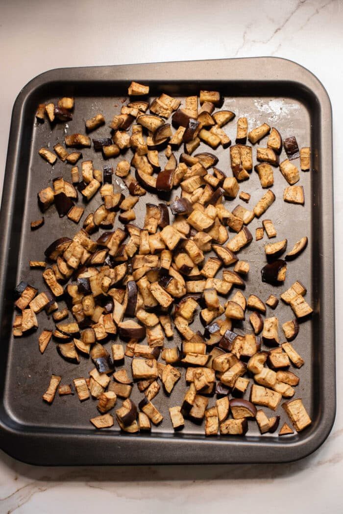 Chopped roasted eggplant pieces spread on a dark baking sheet hint at the solid start to an irresistible eggplant casserole or your next favorite eggplant recipe.