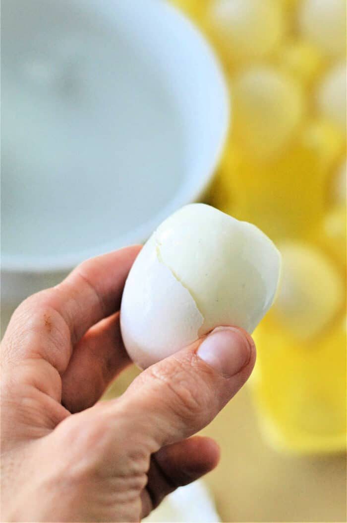 A hand holding a partially peeled boiled egg. In the background, there is a white bowl and a yellow egg carton containing several boiled eggs, hinting they might have been cooked using an eggs in toaster oven method.