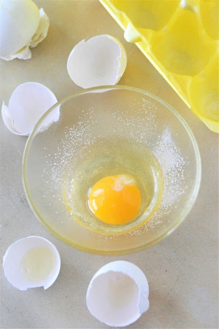 A clear glass bowl with uncooked egg yolk and white on a light surface. Surrounding it are broken eggshells, a yellow egg carton, and a note about using eggs in a toaster oven for quick breakfasts.