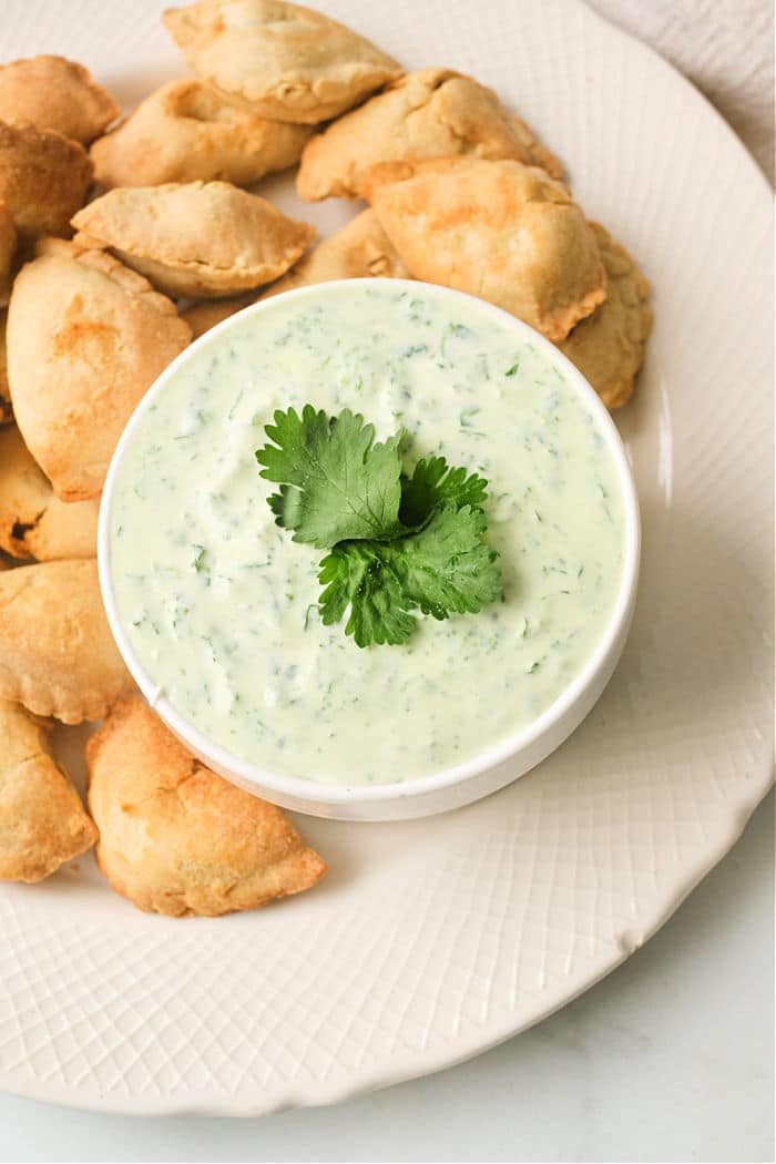 A bowl of creamy green empanada dipping sauce garnished with cilantro sits at the center, surrounded by baked empanadas on a white plate.