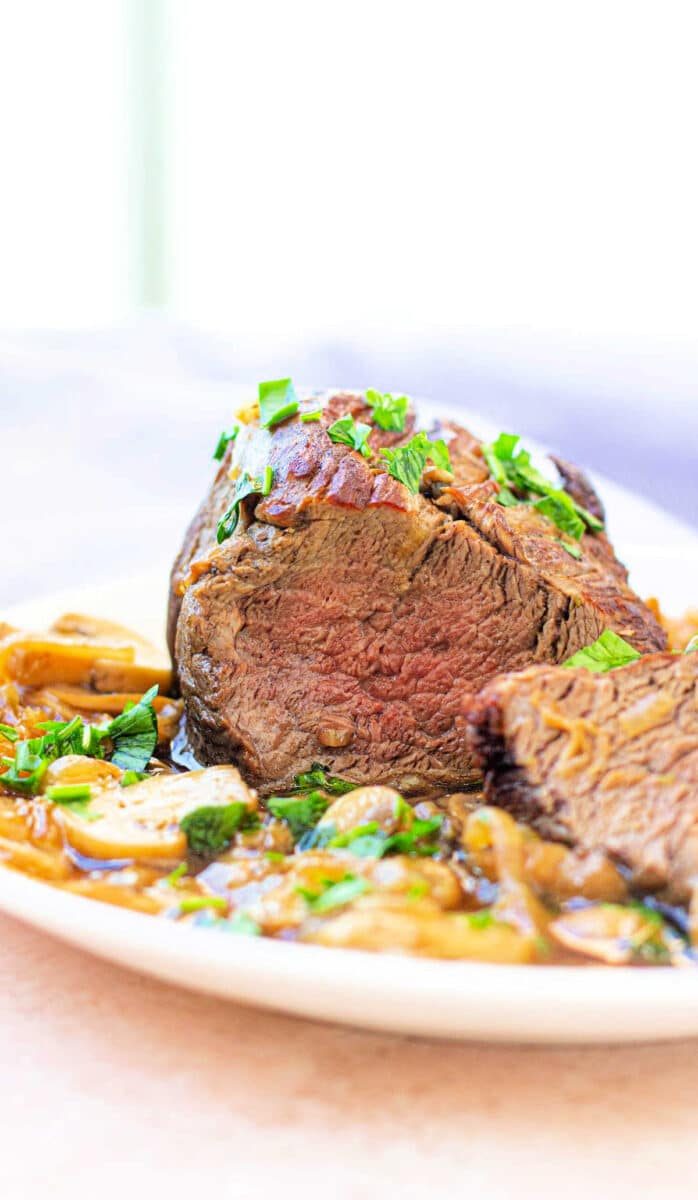 Sliced eye of round roast perfectly prepared garnished with herbs and served alongside sautéed mushrooms and onions.