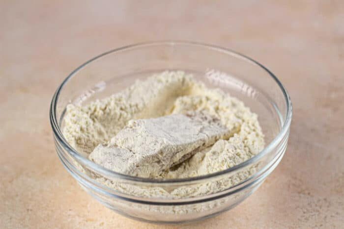 A glass bowl sits on a light-colored surface, holding pieces of raw fish coated in flour, reminiscent of a classic fish fry preparation.