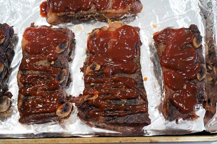Three pieces of barbecued Flanken Short Ribs topped with sauce on a foil-lined baking sheet.