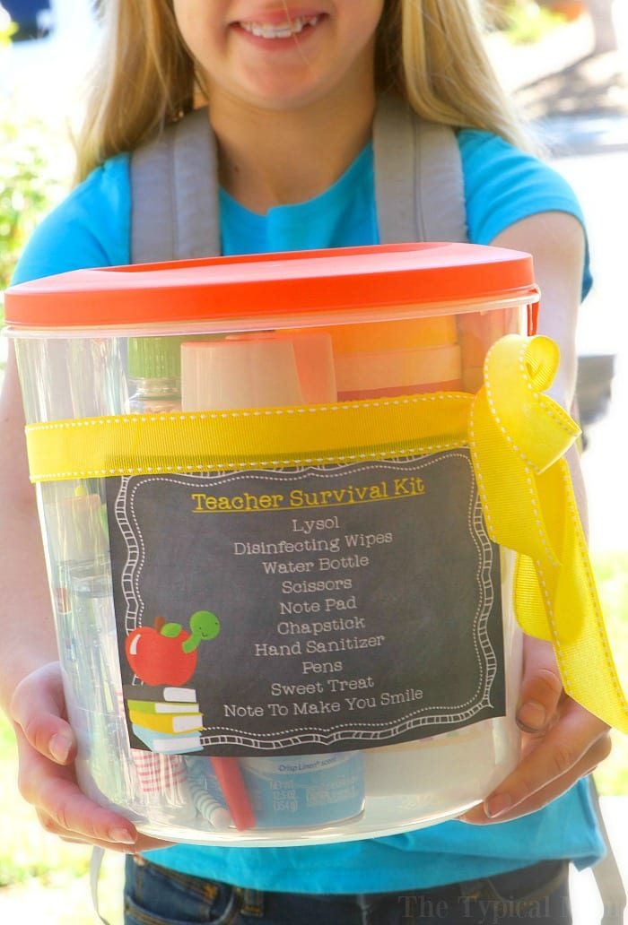 A person in a blue shirt holds a clear container labeled Back to School Teacher Survival Kit, wrapped with a yellow ribbon. Contents listed include disinfecting wipes, water bottle, scissors, note pad, chapstick, hand sanitizer, pens, sweet treat, and a note.