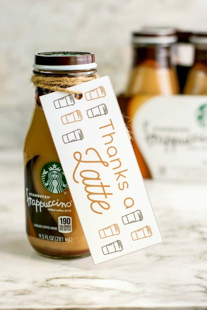 A bottled Starbucks Frappuccino with a label tied around its neck. The label reads "Thanks a Latte" with charming coffee cup illustrations, making it a solid appreciation gift idea. A blurred background features more bottles, adding to the delightful display.