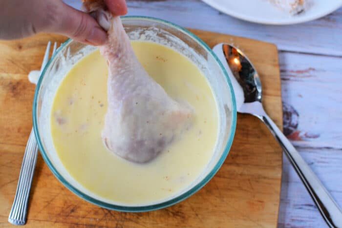Hand dipping raw chicken legs into creamy batter in a glass bowl on a wooden board, ready to be transformed into good pan fried chicken legs, with a spoon and fork beside it.