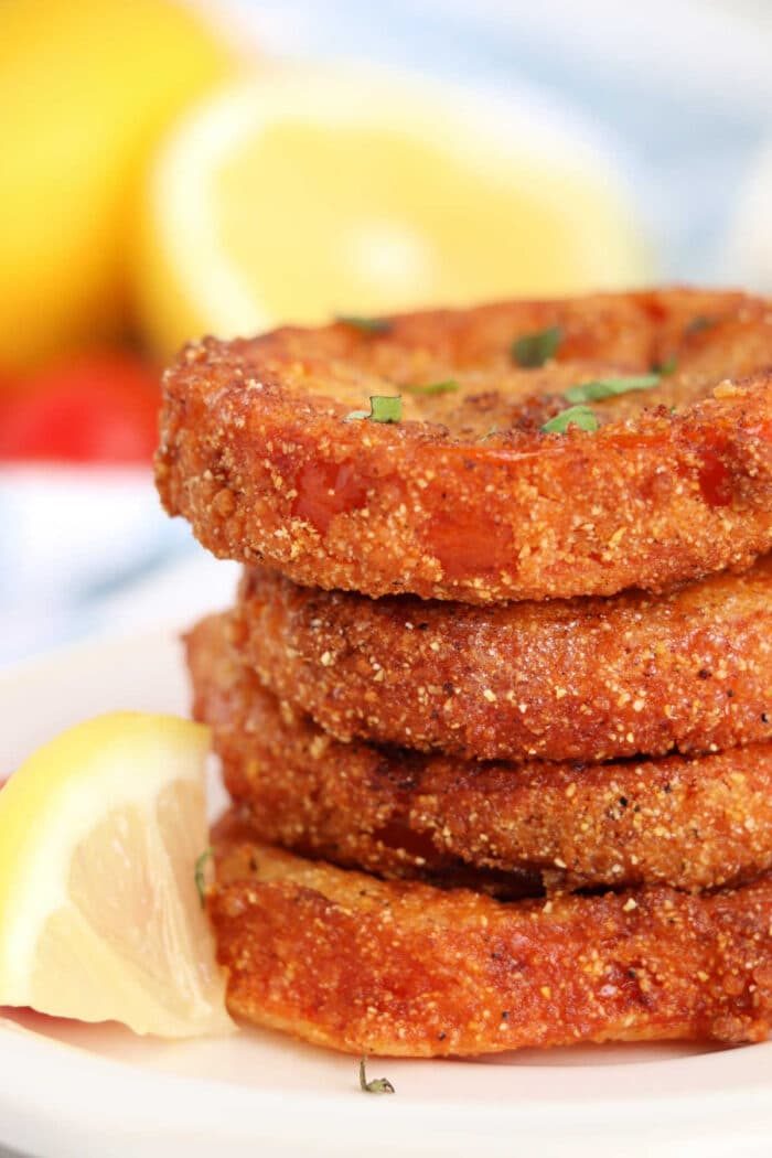 Stacked crispy fried red tomatoes are garnished with fresh herbs, served alongside lemon wedges in the background.