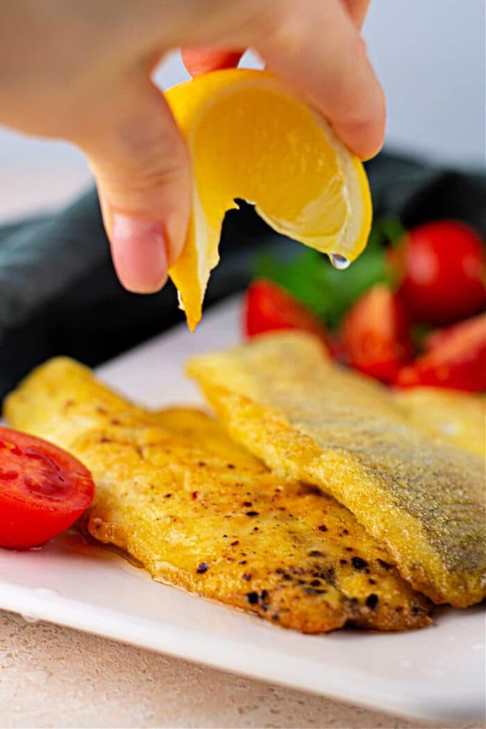 Hand squeezing lemon over seasoned fried Swai fish with cherry tomatoes on a white plate.