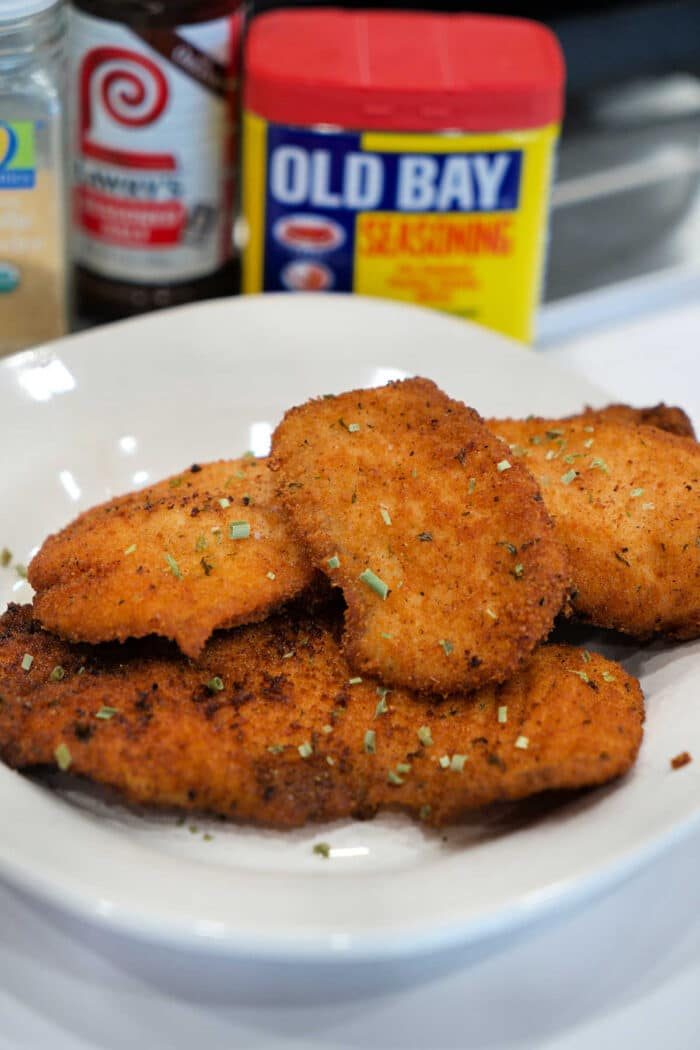 A plate of golden, breaded fried chicken cutlets, garnished with herbs, with seasoning containers behind—solid for those who love a great Fried Tilapia or classic chicken dish.