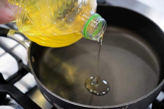 Vegetable oil being poured from a plastic bottle into a black frying pan on a stovetop, ready for preparing a good fried tilapia recipe.