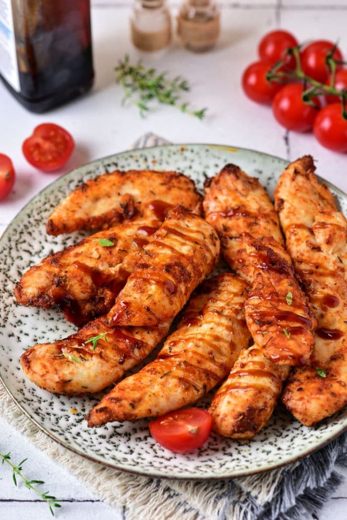 Succulent chicken tenders with a tangy sauce are elegantly plated, garnished with fresh herbs and vibrant cherry tomatoes, effortlessly prepared from frozen chicken tenderloins in an air fryer for ultimate convenience and taste.