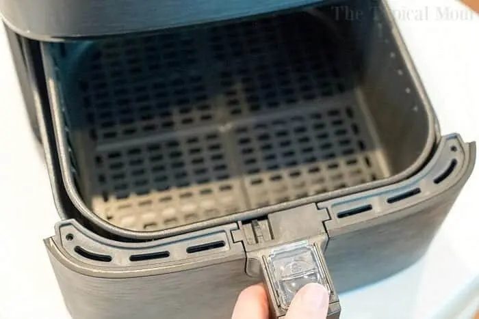 A close-up photo of a hand pulling out the basket of an open air fryer on a light-colored surface. The interior of the air fryer basket is empty and features a mesh design, solid for cooking your favorite dishes like bang bang shrimp. The edge of the air fryer has a latch mechanism for securing the basket.