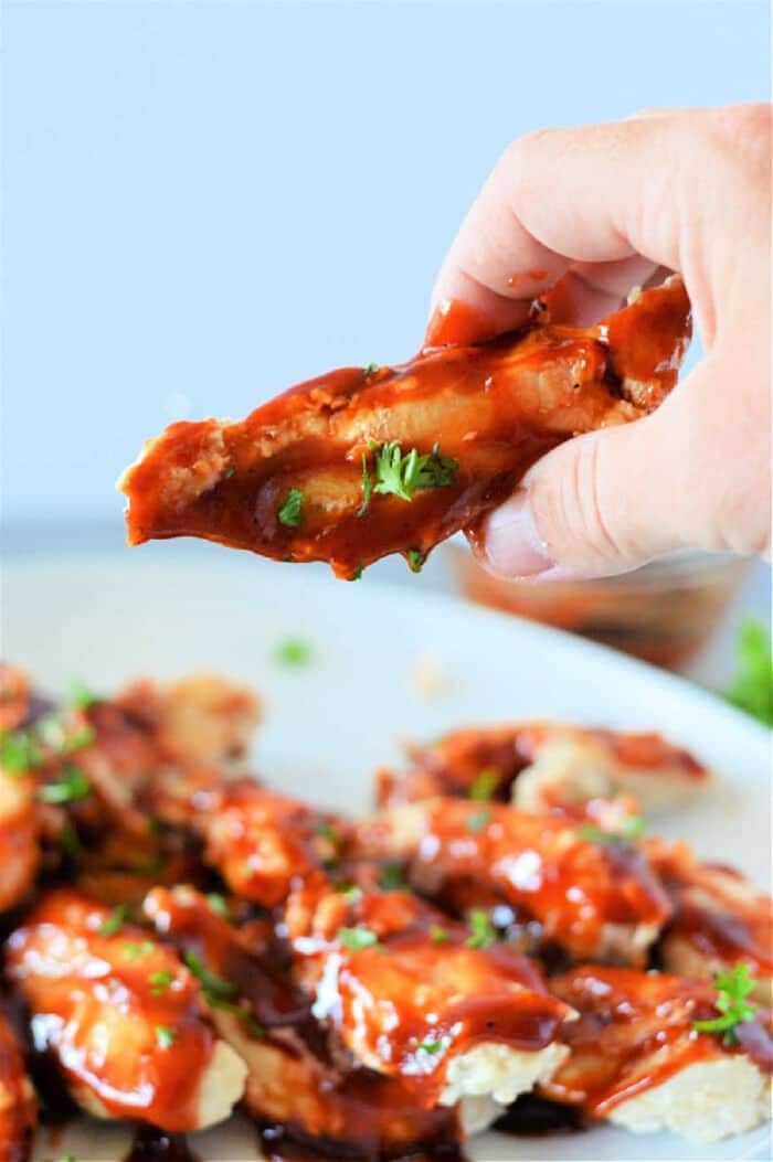 A hand holds a saucy chicken wing above a plate of barbecue wings, garnished with chopped herbs—solid inspiration for making frozen chicken tenders in the Instant Pot at home.