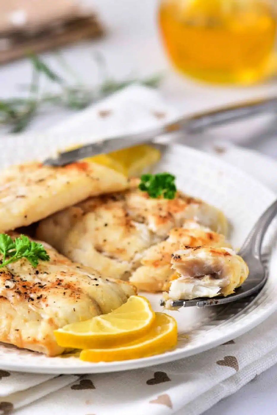 A white plate features air-fried fish fillets garnished with lemon slices and a sprig of parsley. A fork holds a piece of the flaky delight, while a blurred glass with a yellow drink sits in the background. The tablecloth is white with heart patterns, emphasizing this good twist on frozen cod.