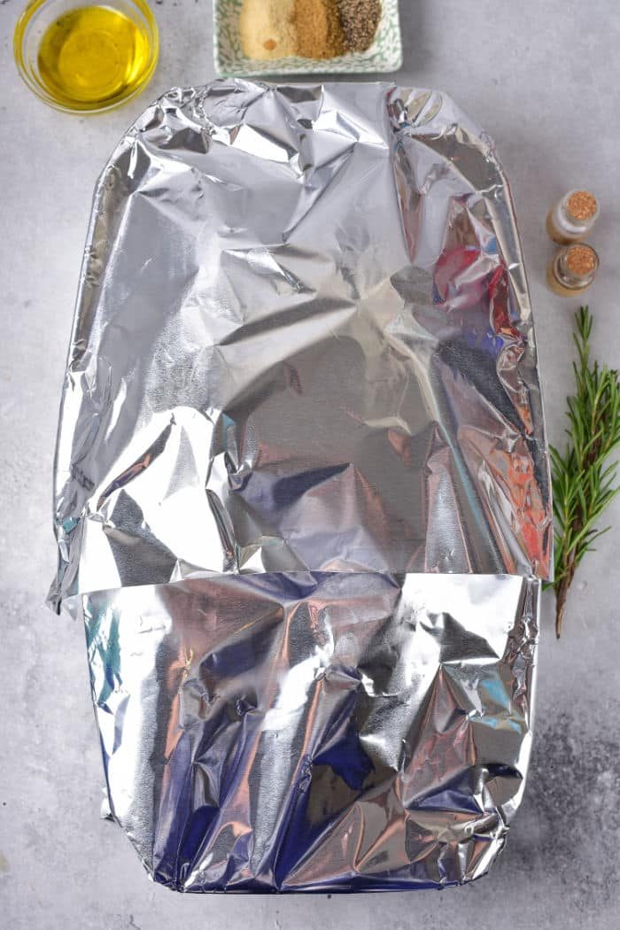 A frozen duck inside a baking dish, which is covered with aluminum foil and centered on the marble countertop. Nearby are a small bowl of olive oil, a plate with seasoning, two small shakers, and a sprig of rosemary.