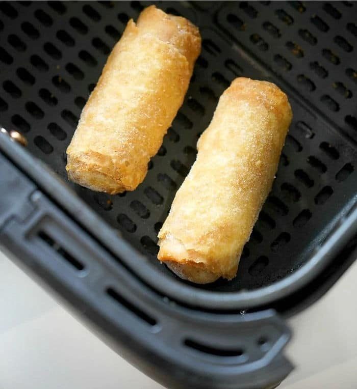 Two frozen egg rolls rest in an air fryer basket, their crispy shells hinting at the convenience of preparing frozen egg rolls in an air fryer.
