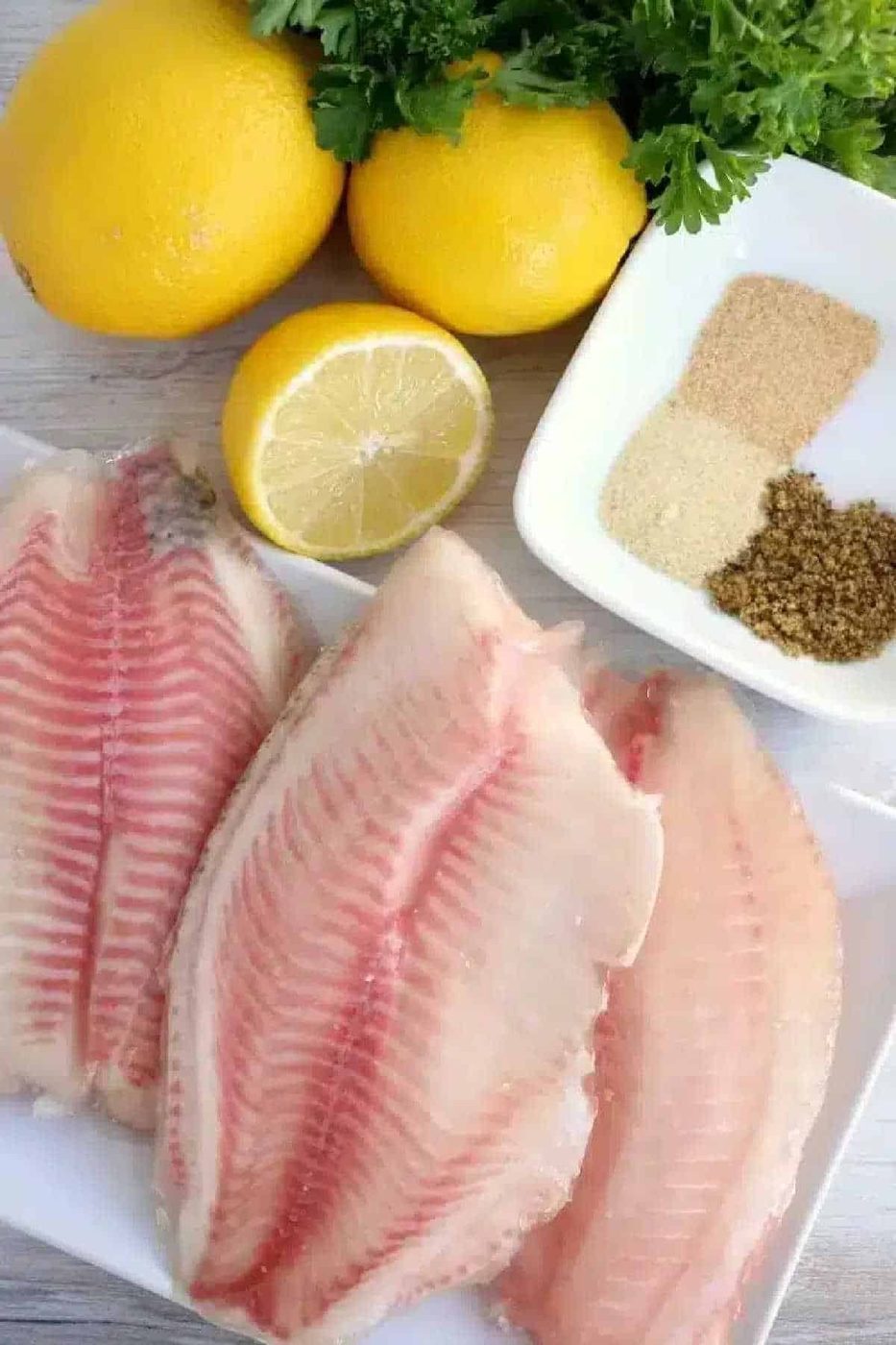Three raw fish fillets, lemons, fresh herbs, and a dish with three kinds of spices on a table—solid ingredients if you're looking to try frozen fish in air fryer for a quick and good meal.