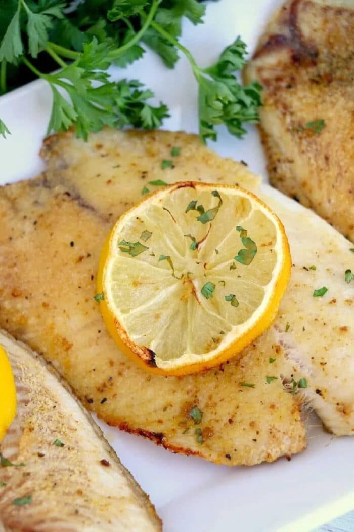 Seasoned frozen fish in air fryer fillet garnished with parsley and a slice of lemon, served on a white plate.