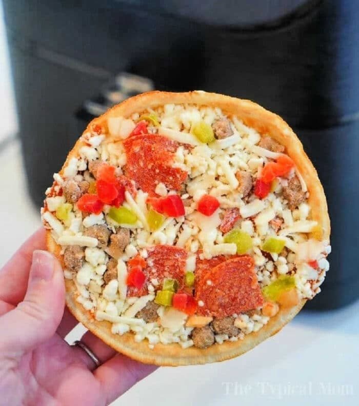 A hand holds a small, round frozen pizza, topped with diced green and red bell peppers, pepperoni slices, sausage crumbles, and shredded cheese. In the background is a black appliance—solid for cooking frozen pizza in an air fryer.
