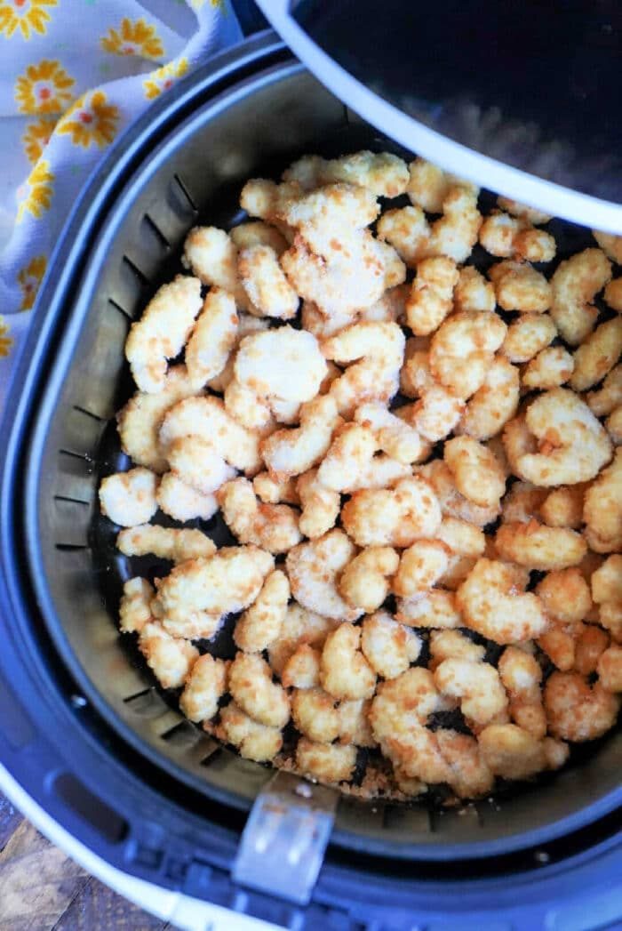 An air fryer basket brimming with crispy, golden-brown popcorn shrimp pieces sits enticingly on a surface, alongside a yellow floral-patterned cloth. It's the solid setup to savor frozen popcorn shrimp in the air fryer next time.