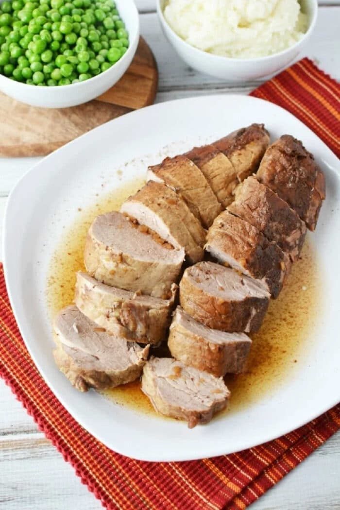 Sliced pork tenderloin cooked from frozen pork loin in Instant Pot, topped with gravy on a white plate, served with mashed potatoes and green peas.