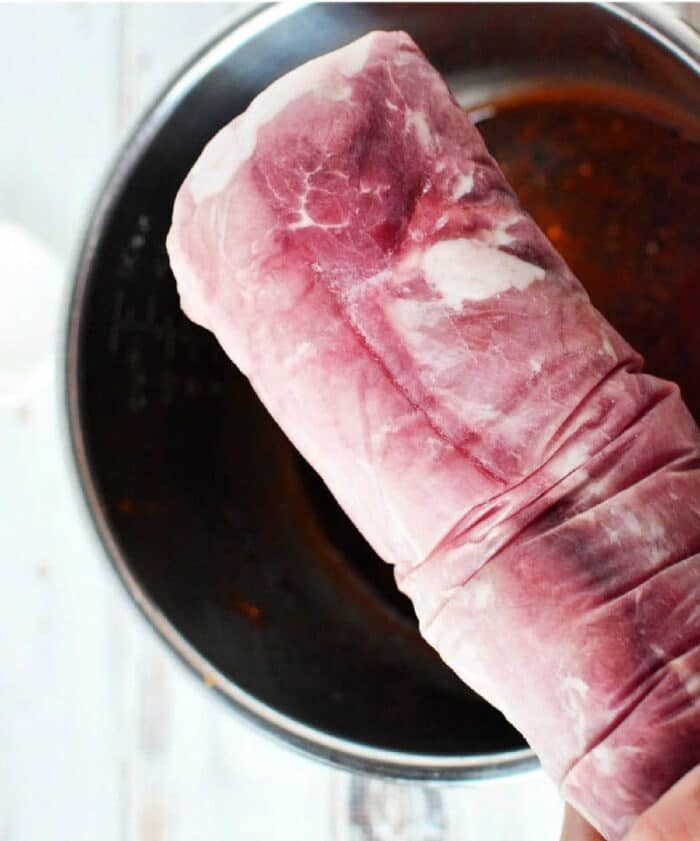 A raw pork tenderloin, solid for making frozen pork loin in an Instant Pot, is held above a metal mixing bowl on a white surface.