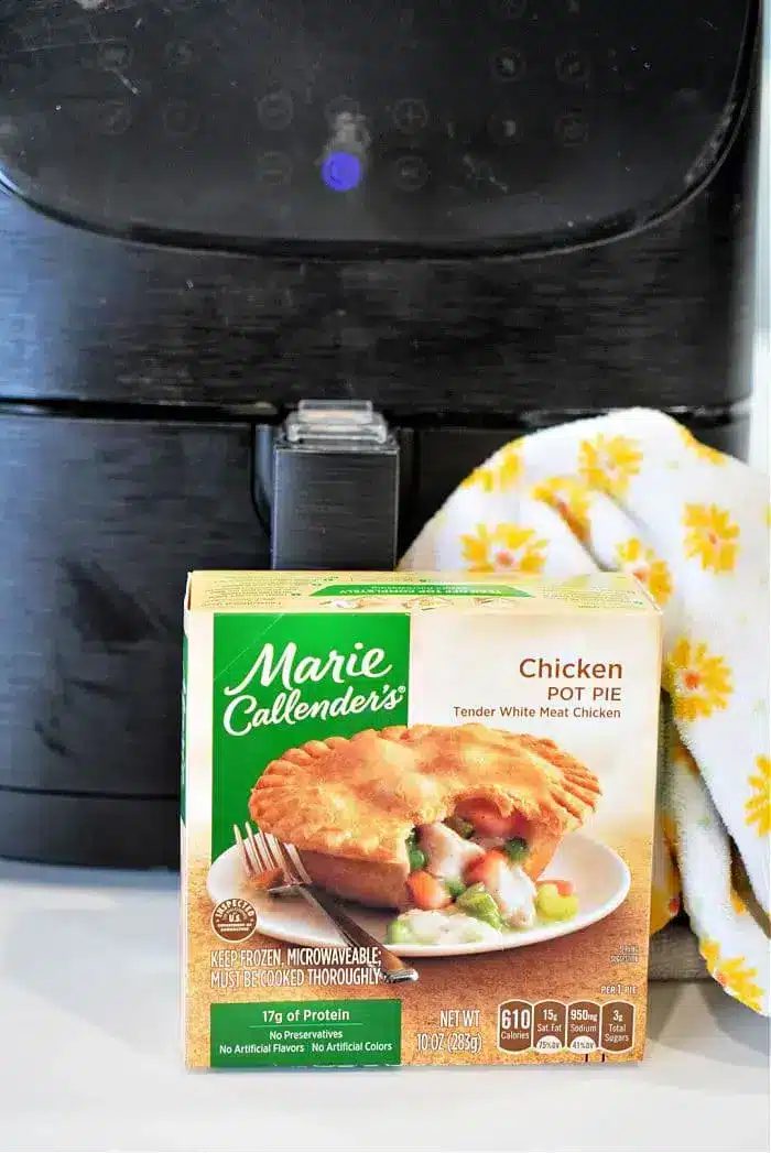 A Marie Callender's frozen pot pie box is placed in front of a black air fryer, ready for a crispy finish. A dish towel with yellow flowers is draped beside it, adding a touch of warmth to the scene.