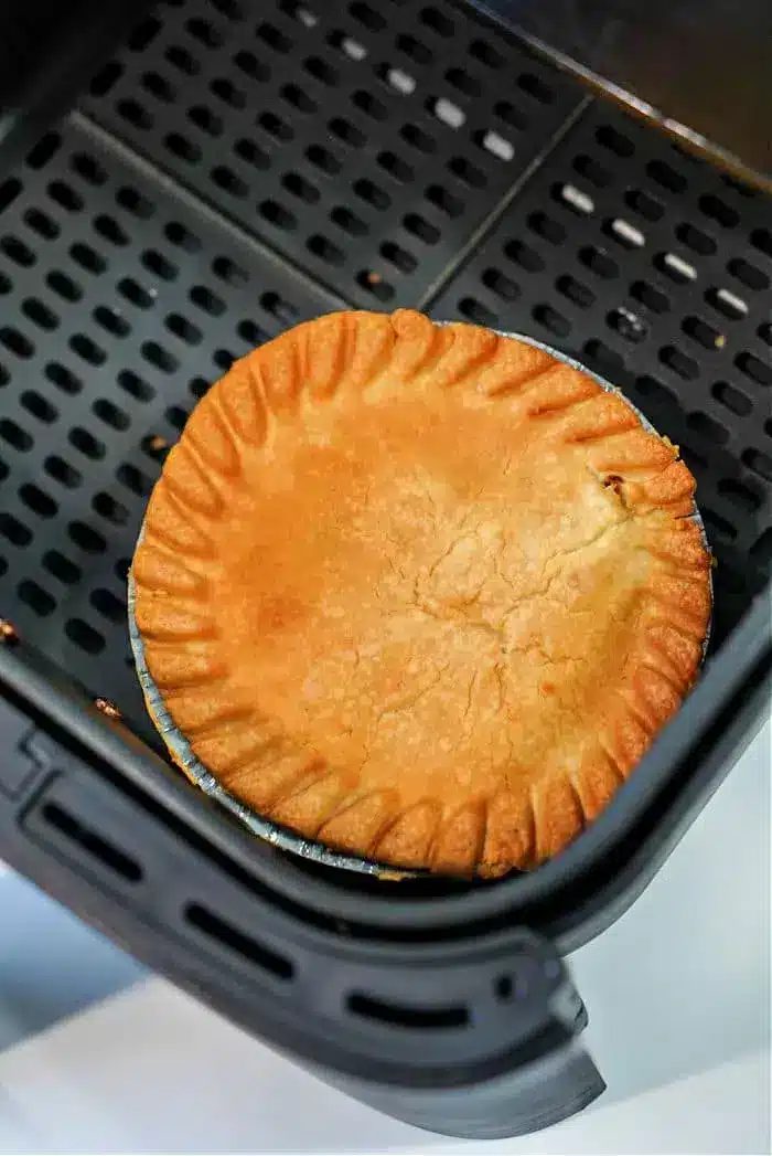 A golden-brown frozen pot pie sits in an air fryer basket, its crust evenly baked with a decorative crimped edge. The air fryer features a perforated base for optimal air circulation, making it solid for transforming frozen delights into crispy perfection.