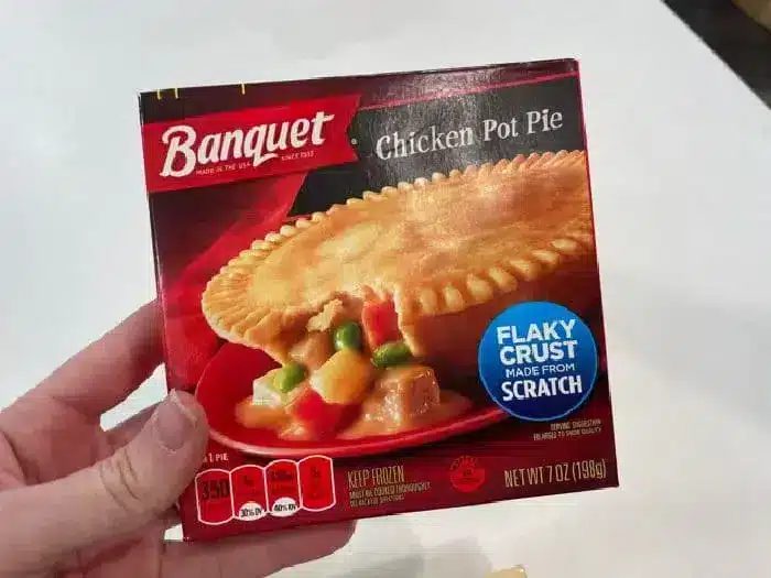 A person's hand is holding a box of Banquet Chicken Pot Pie. The packaging showcases a flaky crust, made from scratch, and suggests cooking the 7-ounce frozen pot pie in an air fryer for optimal crispiness. Nutritional info and instructions are detailed on the box.