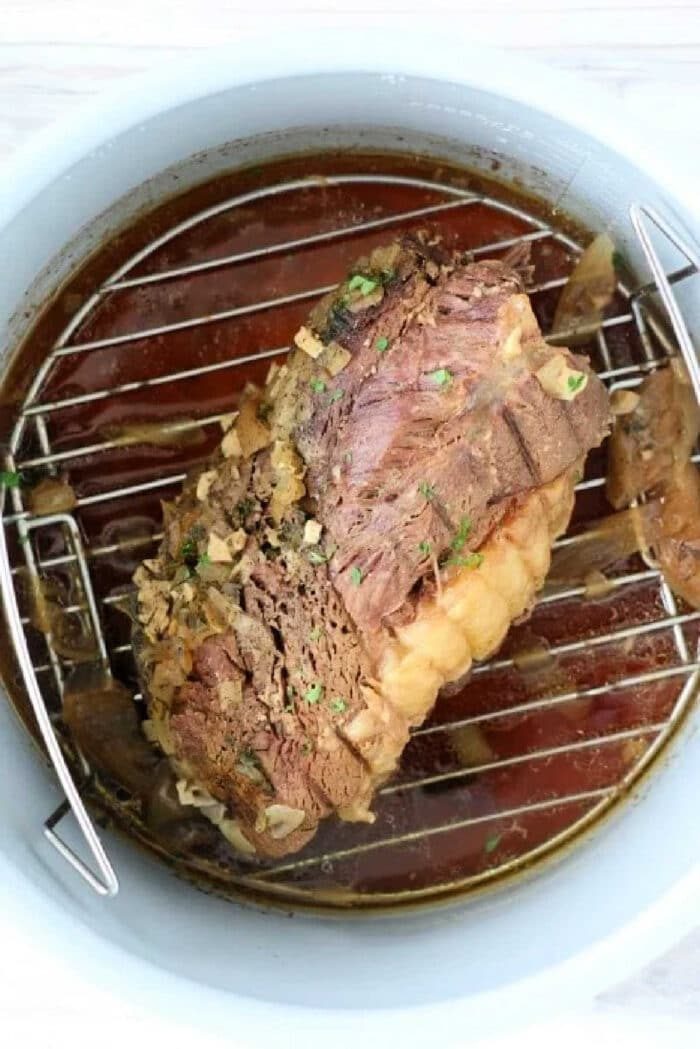 Cooked beef roast topped with herbs and garlic sits on a metal rack inside a pot with broth, solid for making a tender frozen roast in Instant Pot.