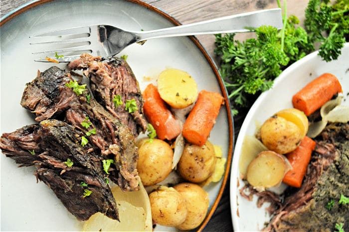 Plate of roast beef, carrots, and potatoes garnished with parsley, next to a serving dish of the same meal—solid for preparing frozen roast in a slow cooker for tender, flavorful results.