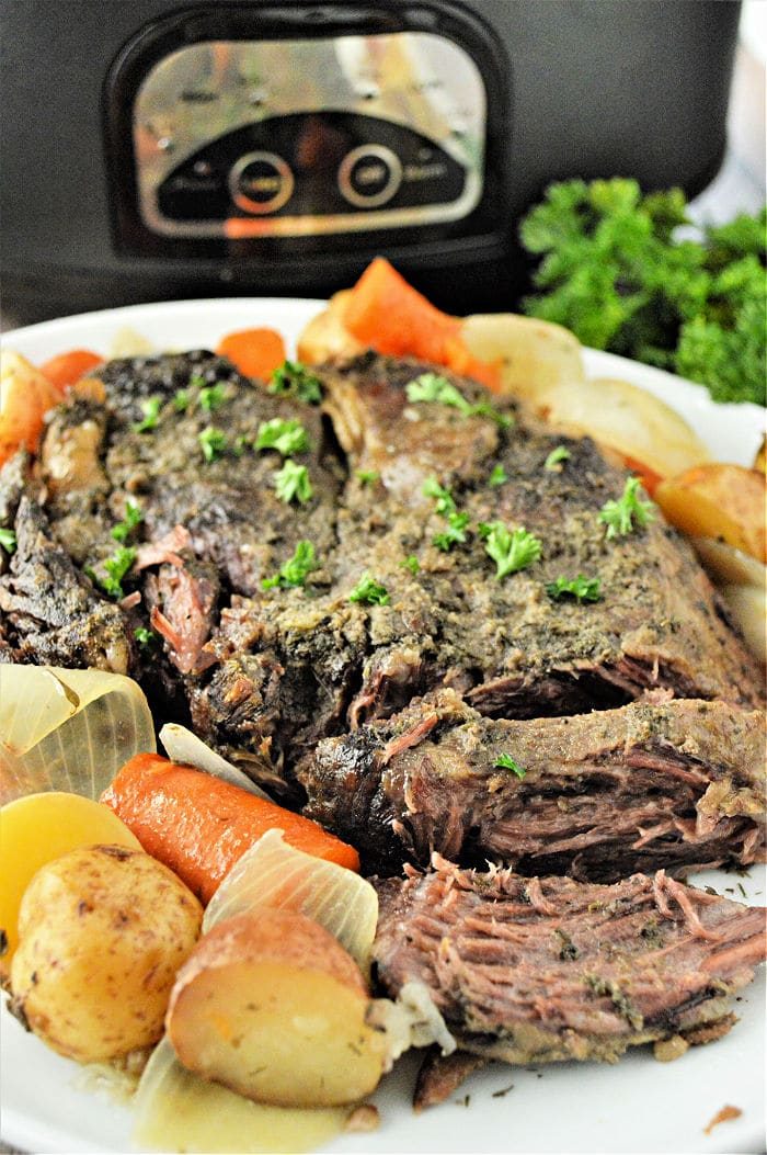 Tender pot roast with carrots, potatoes, and onions on a white plate in front of a slow cooker—solid for making even a frozen roast in the slow cooker.