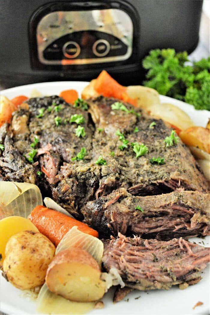 A plate of pot roast with potatoes, carrots, and onions, garnished with parsley, sits in front of a slow cooker—solid for making a frozen roast in slow cooker for a hearty meal.