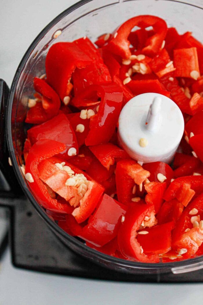 A food processor filled with cut red bell peppers, seeds, and a hint of garlic, ready for blending into a good chili paste recipe.