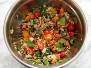 A metal bowl filled with mixed vegetables, cherry tomatoes, rice, and slices of grilled stuffed peppers sits on a light marble surface.
