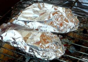 Two foil-wrapped food items, possibly grilled stuffed peppers, cook on a grill over hot coals.
