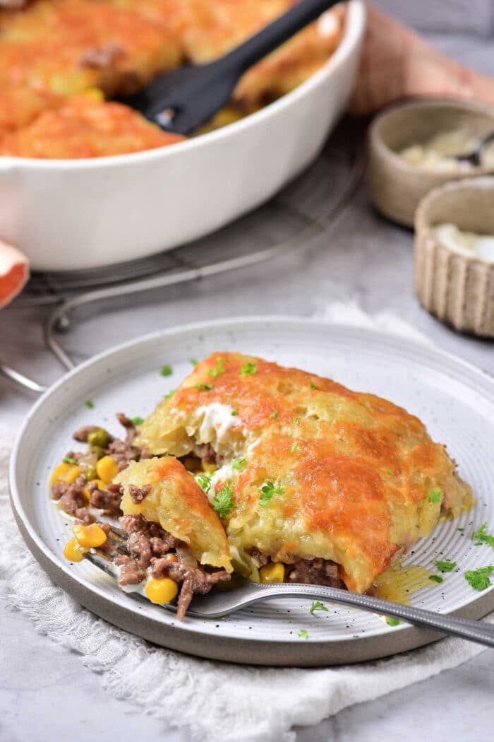 A slice of ground beef casserole with vegetables and melted cheese on top, resembling a ground beef hash brown casserole, with a baking dish in the background.