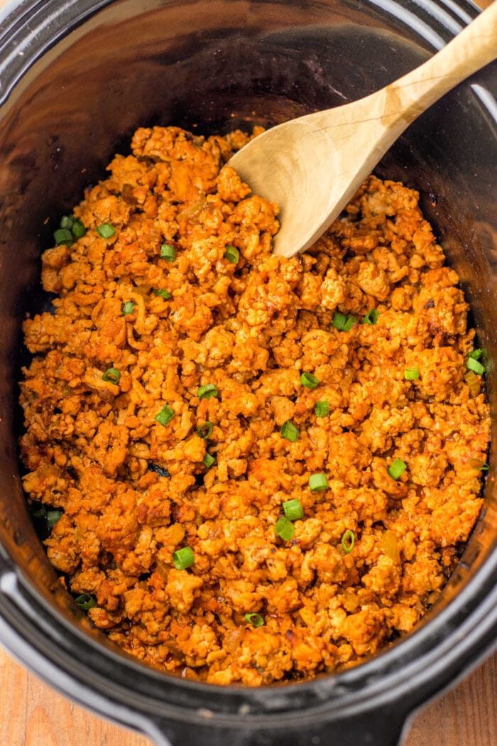 Cooked seasoned ground turkey in a crockpot with a wooden spoon, transforming it into a delightful dish reminiscent of Ground Chicken Sloppy Joes, garnished with chopped green onions.