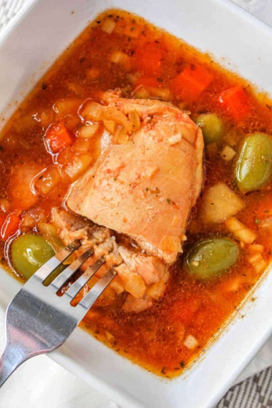 A fork cutting into a piece of cooked chicken in a bowl of Guisado de Pollo, a savory vegetable stew with green olives.