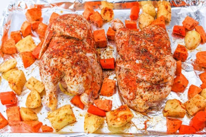 A seasoned half chicken rest amid chunks of sweet potatoes and carrots on a foil-lined baking sheet.