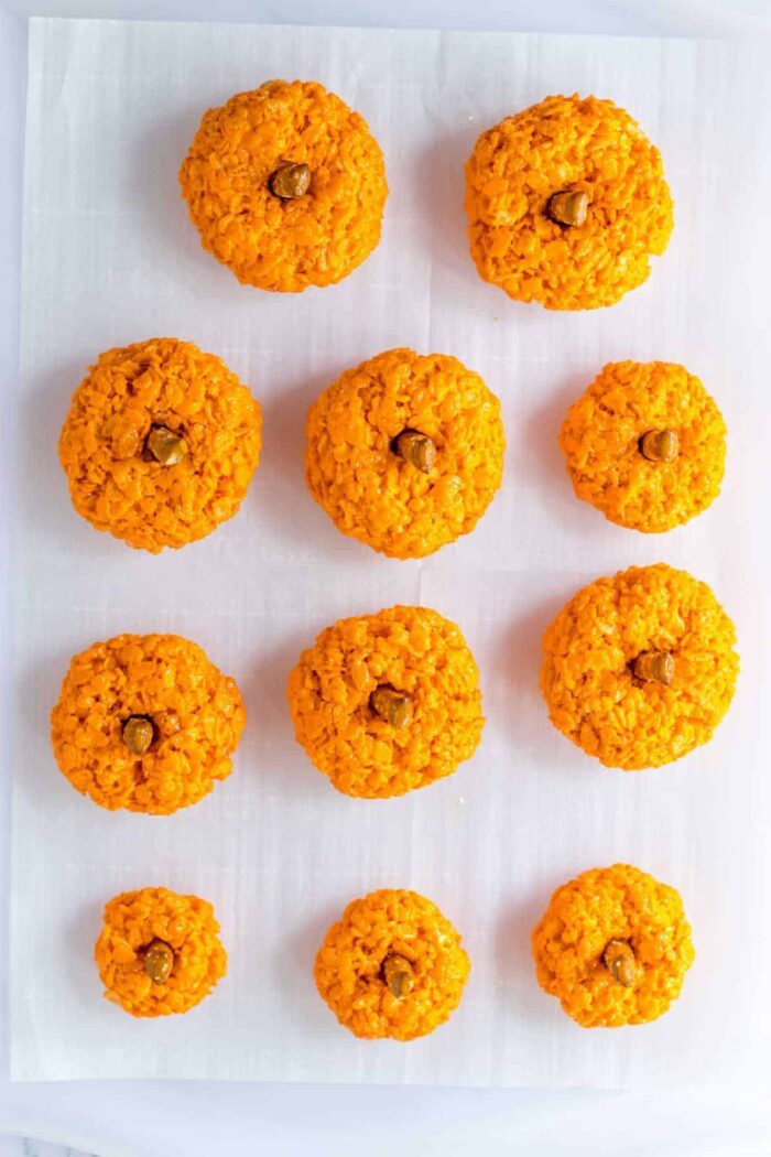 Ten Halloween Rice Krispie Treats shaped like pumpkins with green stems are arranged in rows on white parchment paper.