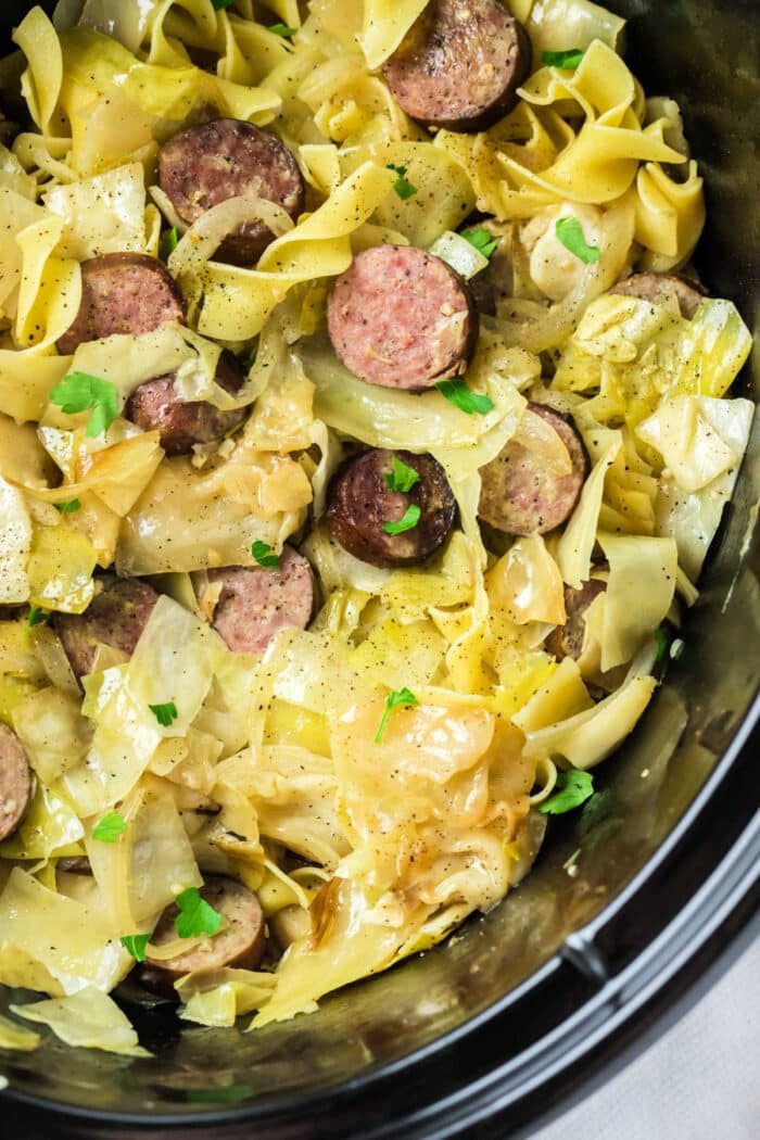 Sliced sausages with cabbage and noodles, reminiscent of a classic Haluski recipe, garnished with chopped parsley in a crockpot.