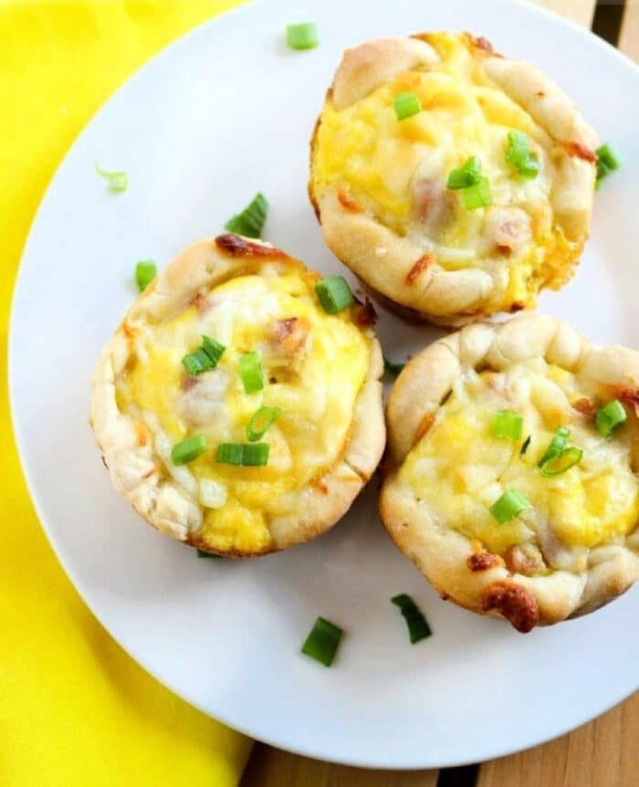 Three baked breakfast muffins filled with cheese, eggs, and green onions are arranged on a white plate. They sit beside a yellow napkin, reminiscent of good ham and cheese biscuits.