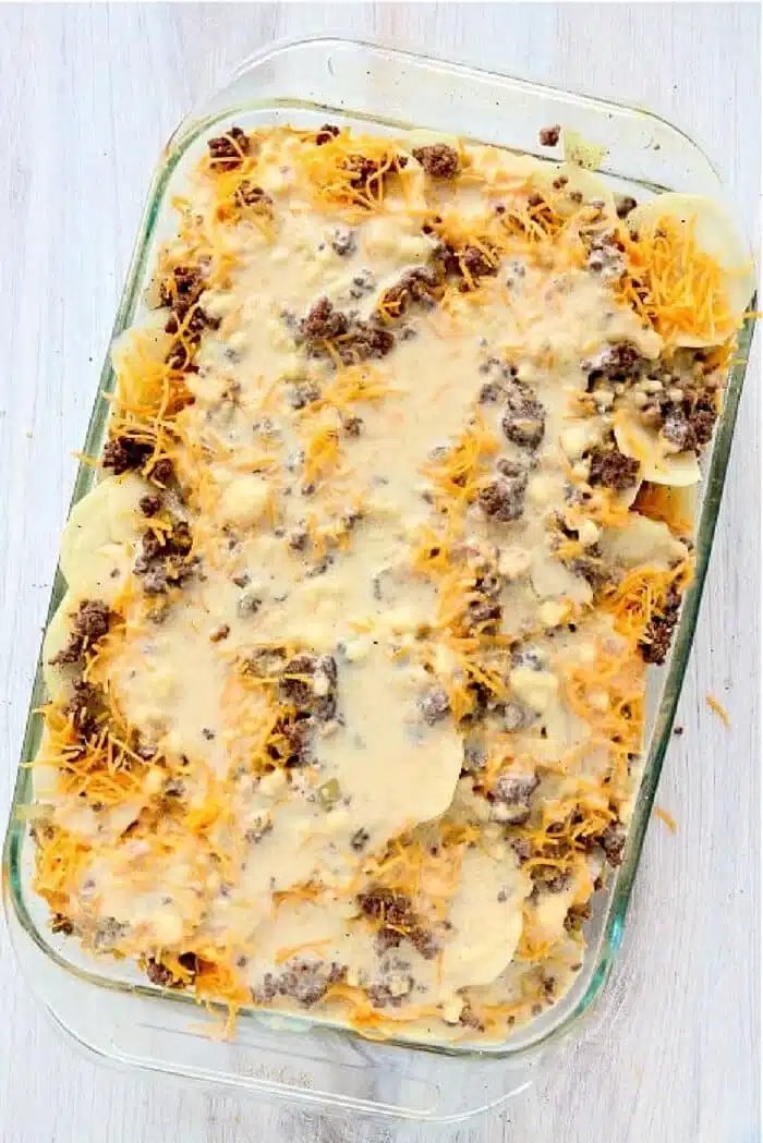 A glass baking dish filled with a good Hamburger HotDish showcases its layered casserole. Visible ingredients include sliced potatoes, browned ground beef, melted cheese, and a creamy sauce. The dish rests on a light-colored wooden surface, inviting you to savor its flavors.